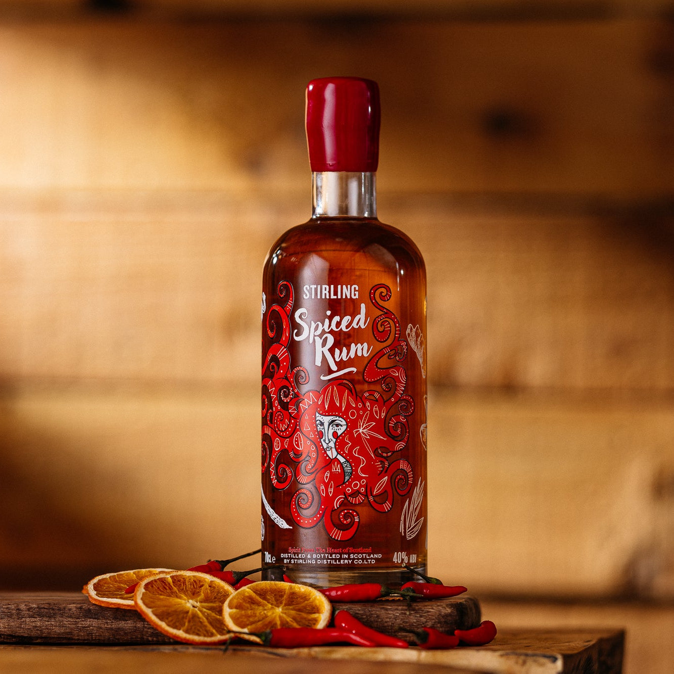A bottle of Stirling Spice Rum on a wooden table with orange and chili peppers around it. Rustic wood panelling background.