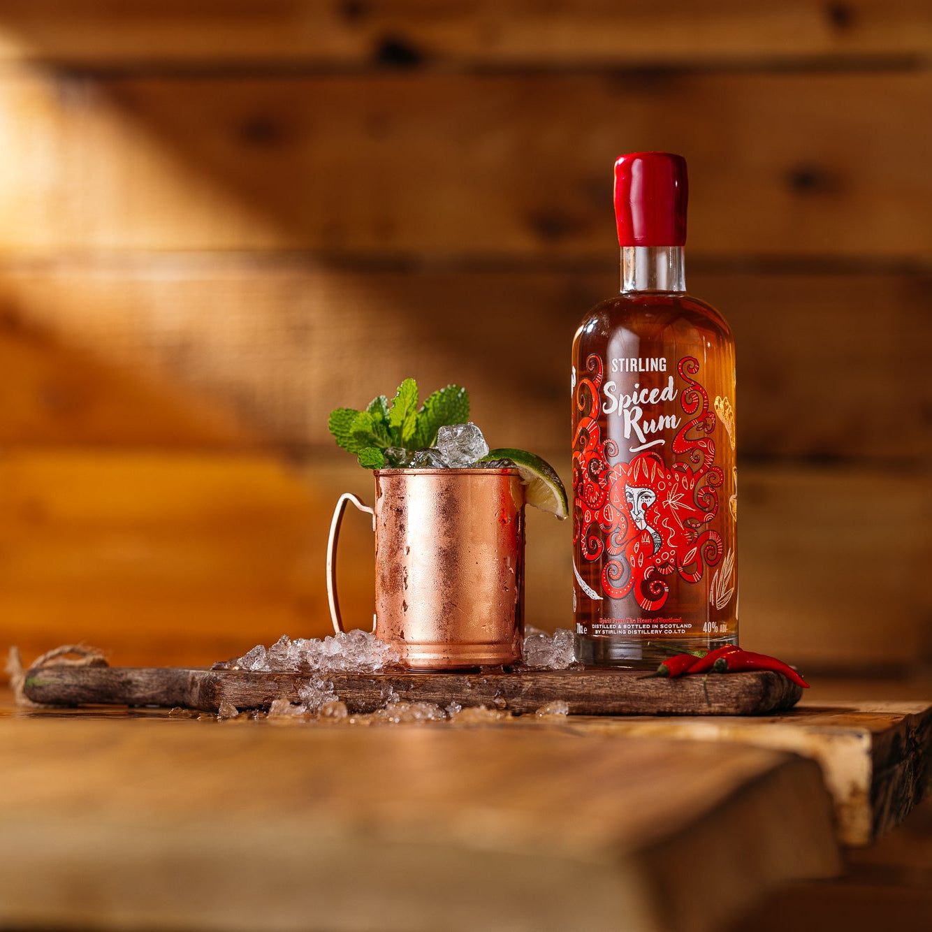A bottle of Stirling Spiced Rum on a wooden table beside a cocktail in a copper mug which is garnished with ice mint and lime. Rustic wood panelling background.