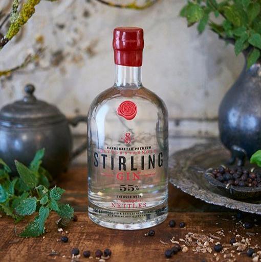 A mini bottle of Battle Strength Gin, red waxed cap with a clear liquid. The label has black and red writing with an illustration of William Wallace on his horse. On a wooden table with the ingredients scattered around the base.