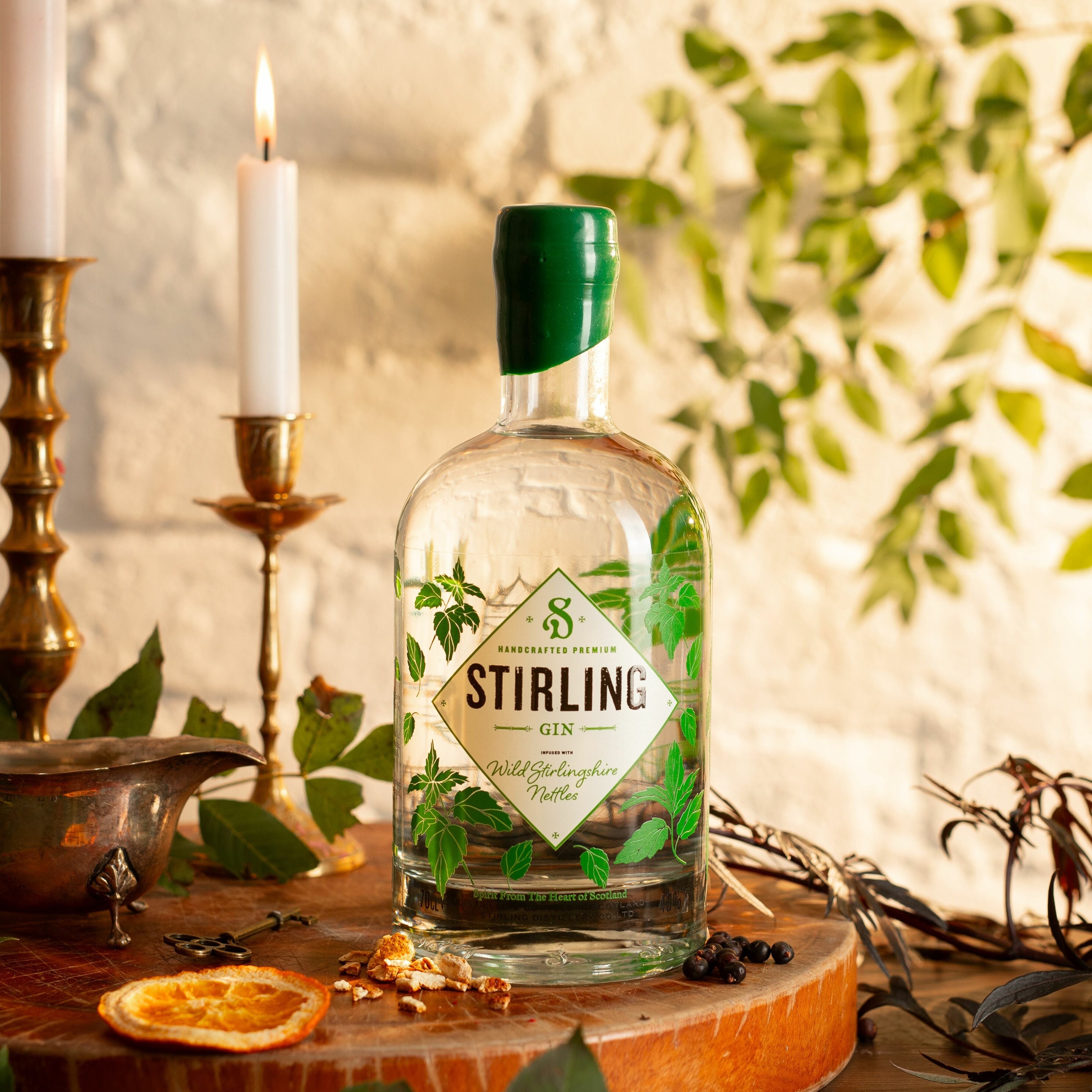 A bottle of Stirling Gin with a green label and wax seal on the lid, decorated with illustrations related to Stirlingshire. On a wooden surface with candles and plants.