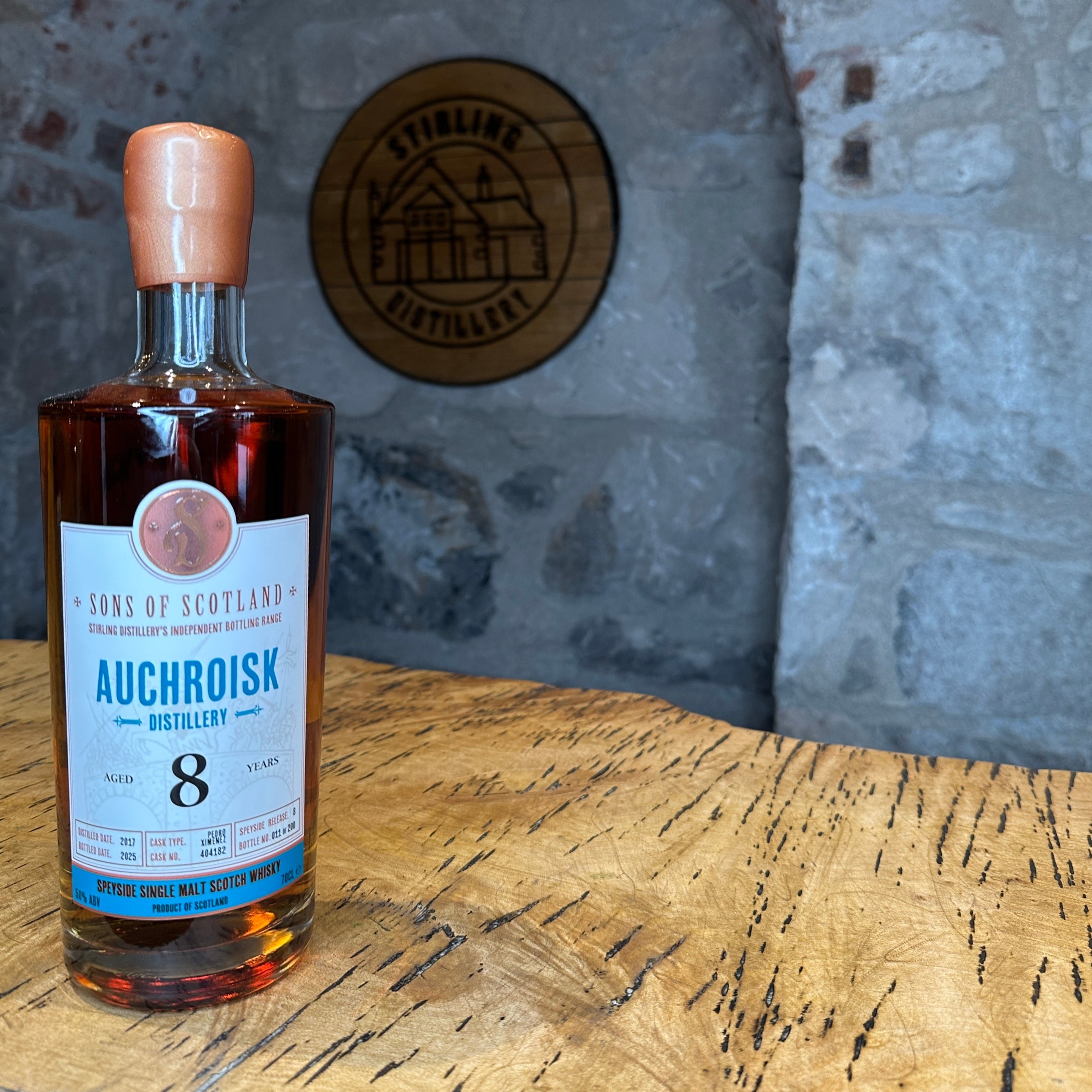 Bottle of Auchroisk whisky on a wooden table with a stone wall background featuring the Stirling Distillery logo