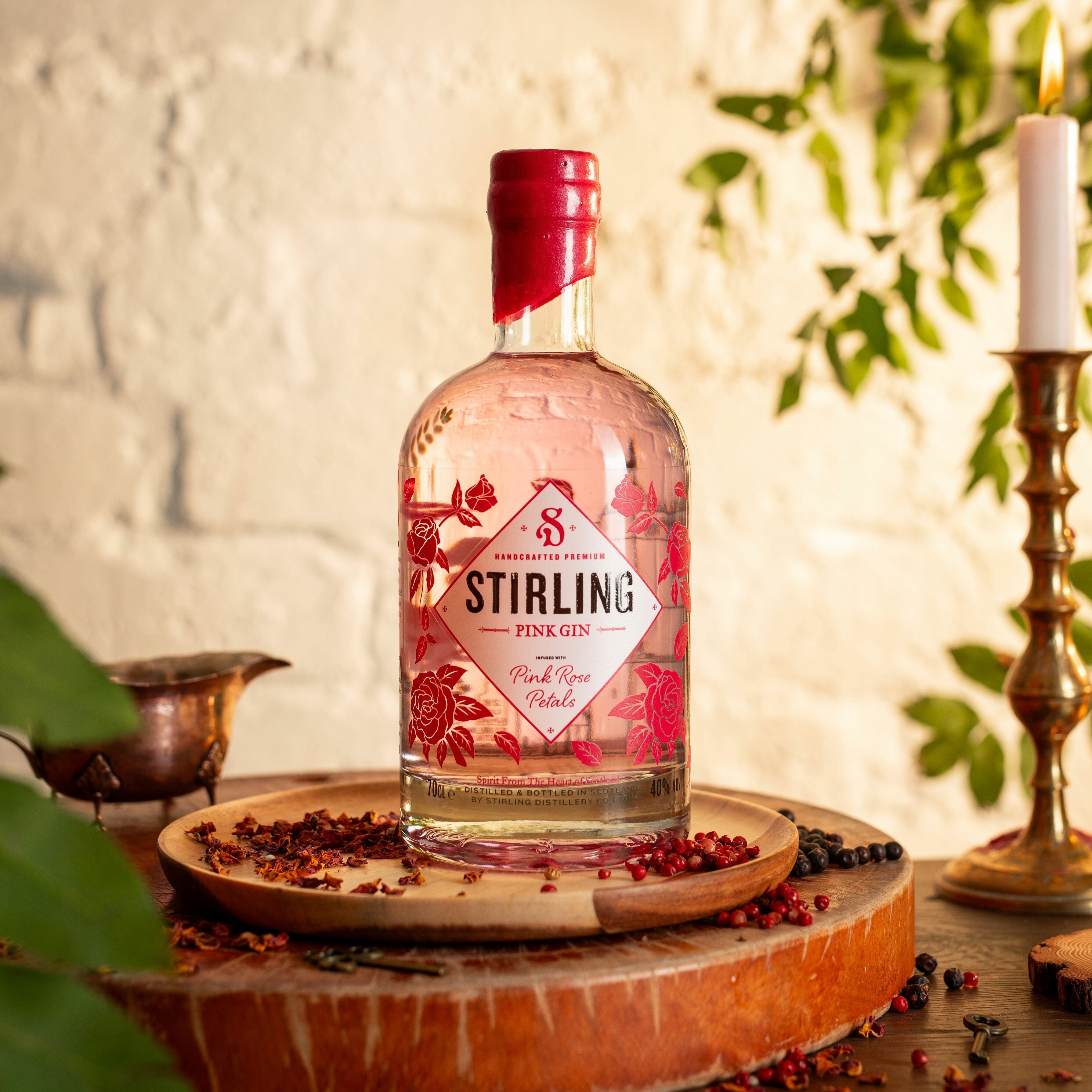 A bottle of Stirling Pink Gin with a pink cap and label featuring illustrations of floral patterns and the ghost of the Pink Lady. Set on a wooden surface with botanics around the base and a stone wall background.
