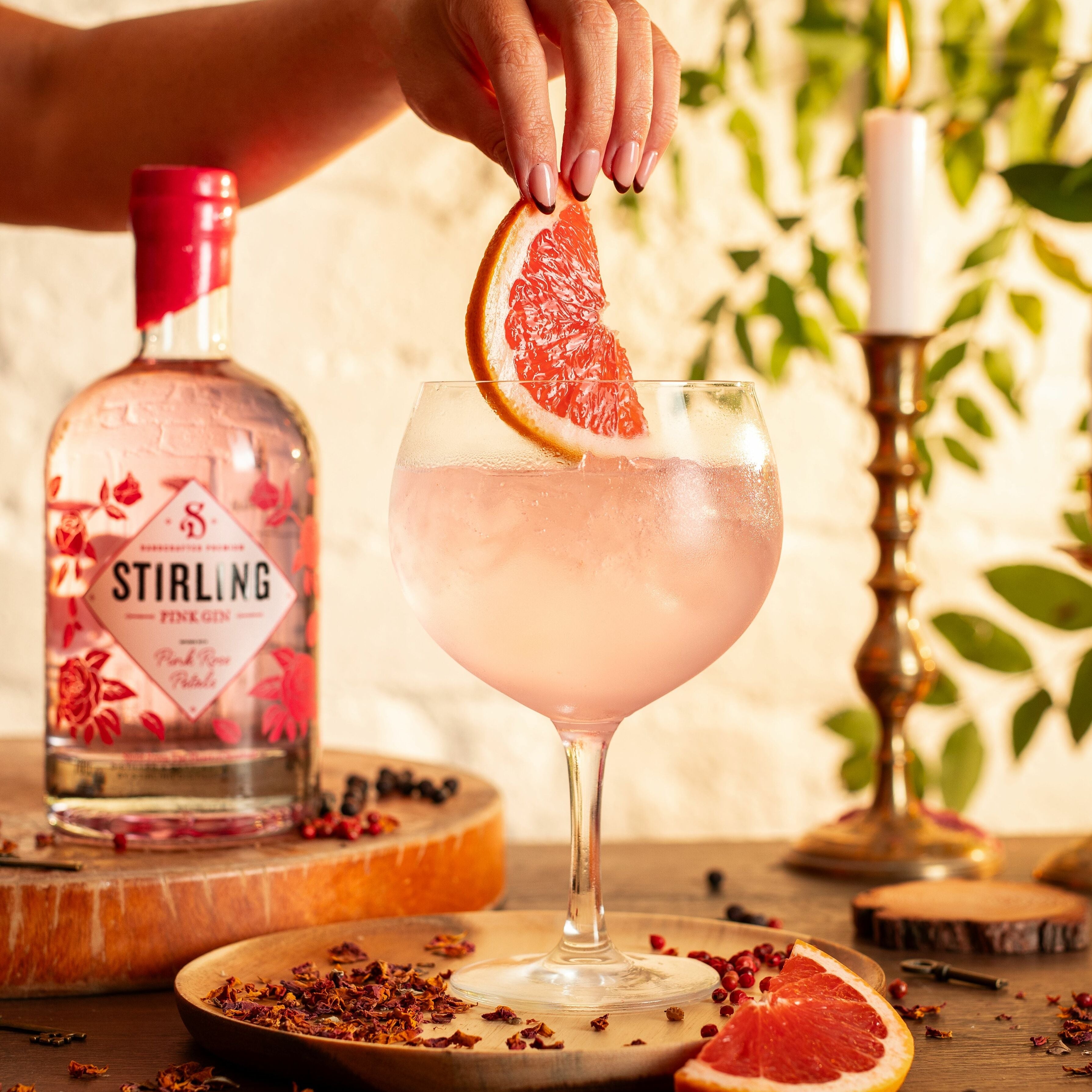 A glass of pink gin and tonic with a hand placing a slice of pink grape fruit into the glass. A bottle of Stirling Pink Gin with a pink cap and label featuring illustrations of floral patterns and the ghost of the Pink Lady is off to the left hand side.