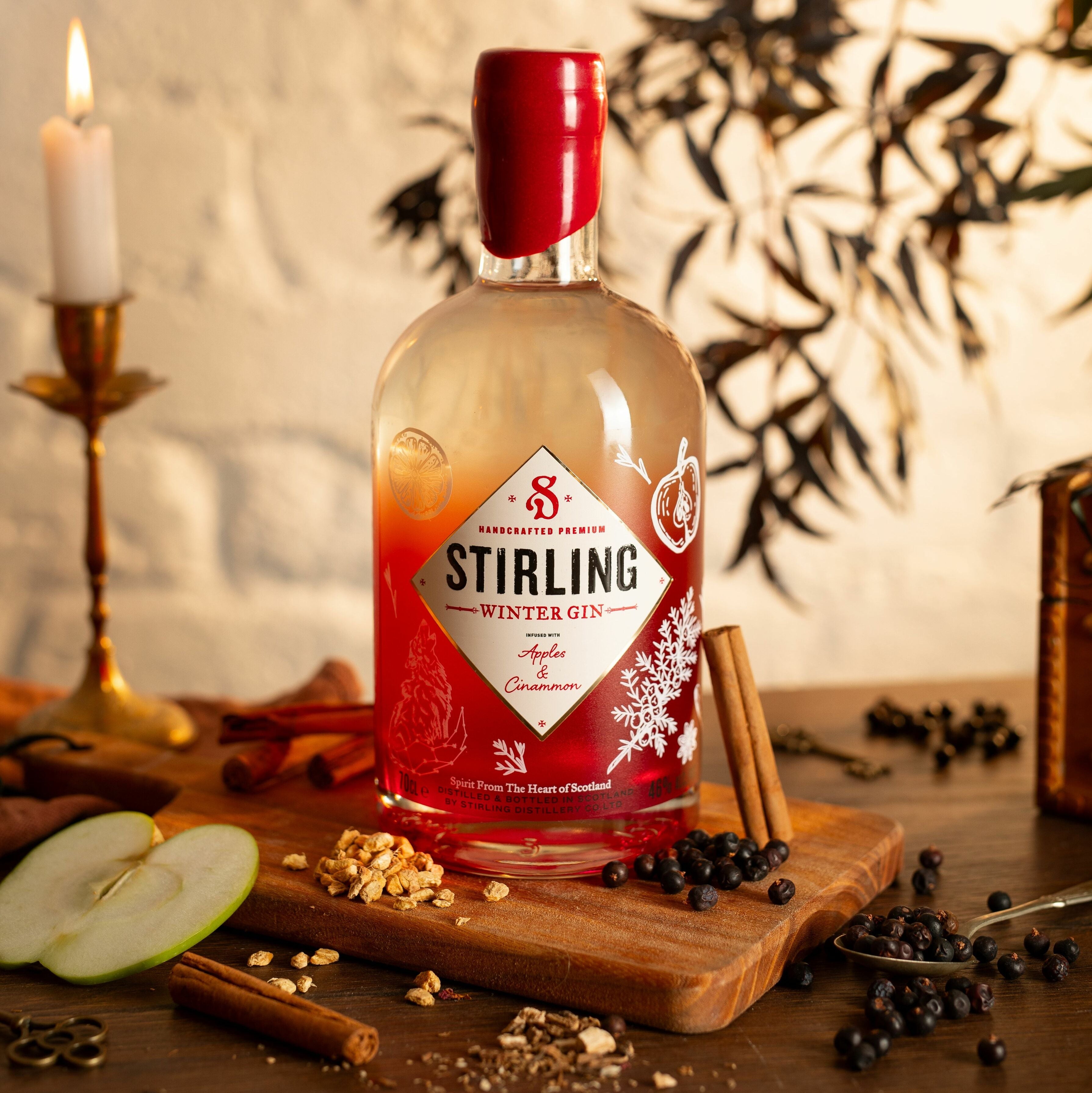 A bottle of Stirling Winter Gin with a red label, featuring illustrations that suggest a festive, wintery theme. On top of a wooden table with the ingredients spread around the bottle. Candle and a plant blurred in the back ground.