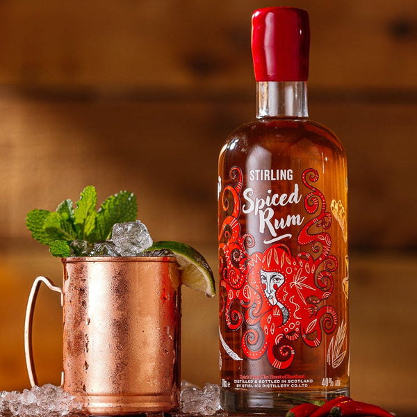 A bottle of Stirling Spiced Rum on a wooden table beside a cocktail in a copper mug which is garnished with ice mint and lime. Rustic wood panelling background.