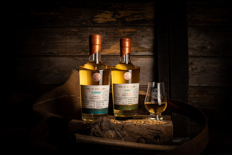 On a wooden surface there are 2 bottles of whisky beside a glencairn glass full of whisky. The table has barley grains on it.
