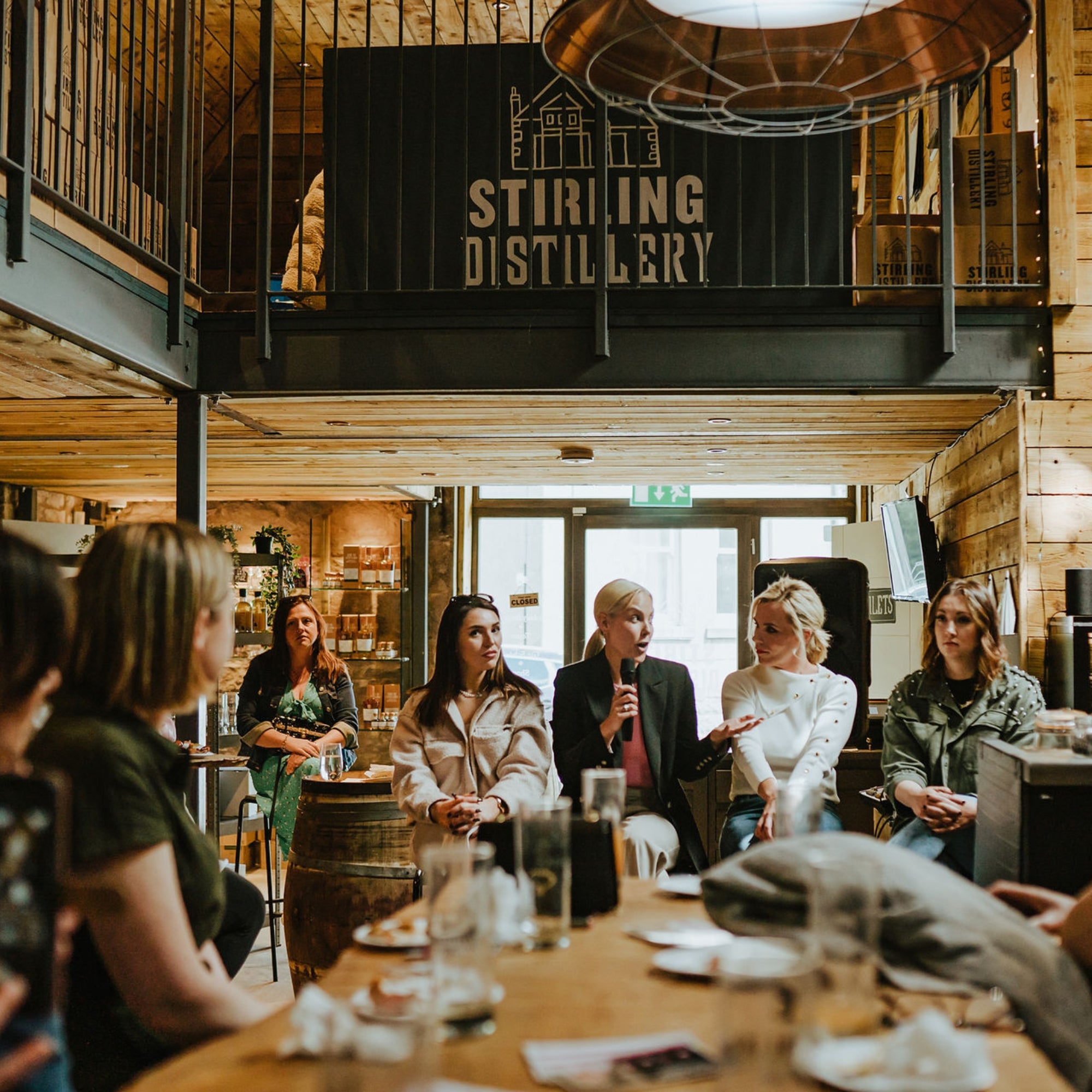 Women are sitting on stools listening to another woman talking on a microphone. They are in the distillery and the Stirling Distillery logo is in the  background.