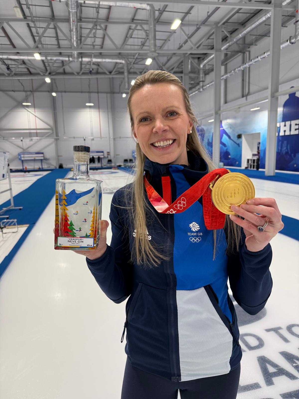 Person holding a gold medal and a box with an Olympic logo in an indoor sports facility.