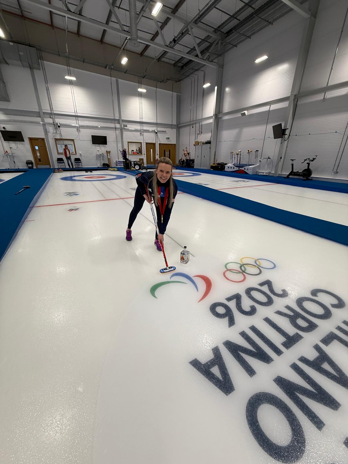 Person curling on an indoor ice rink with 'Cortina 2026' branding.